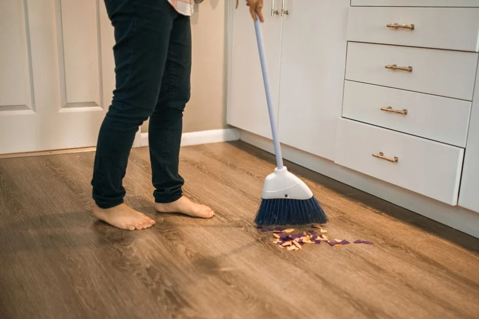 A hand using a feather duster to clean a wooden desk, surrounded by plants and office items.